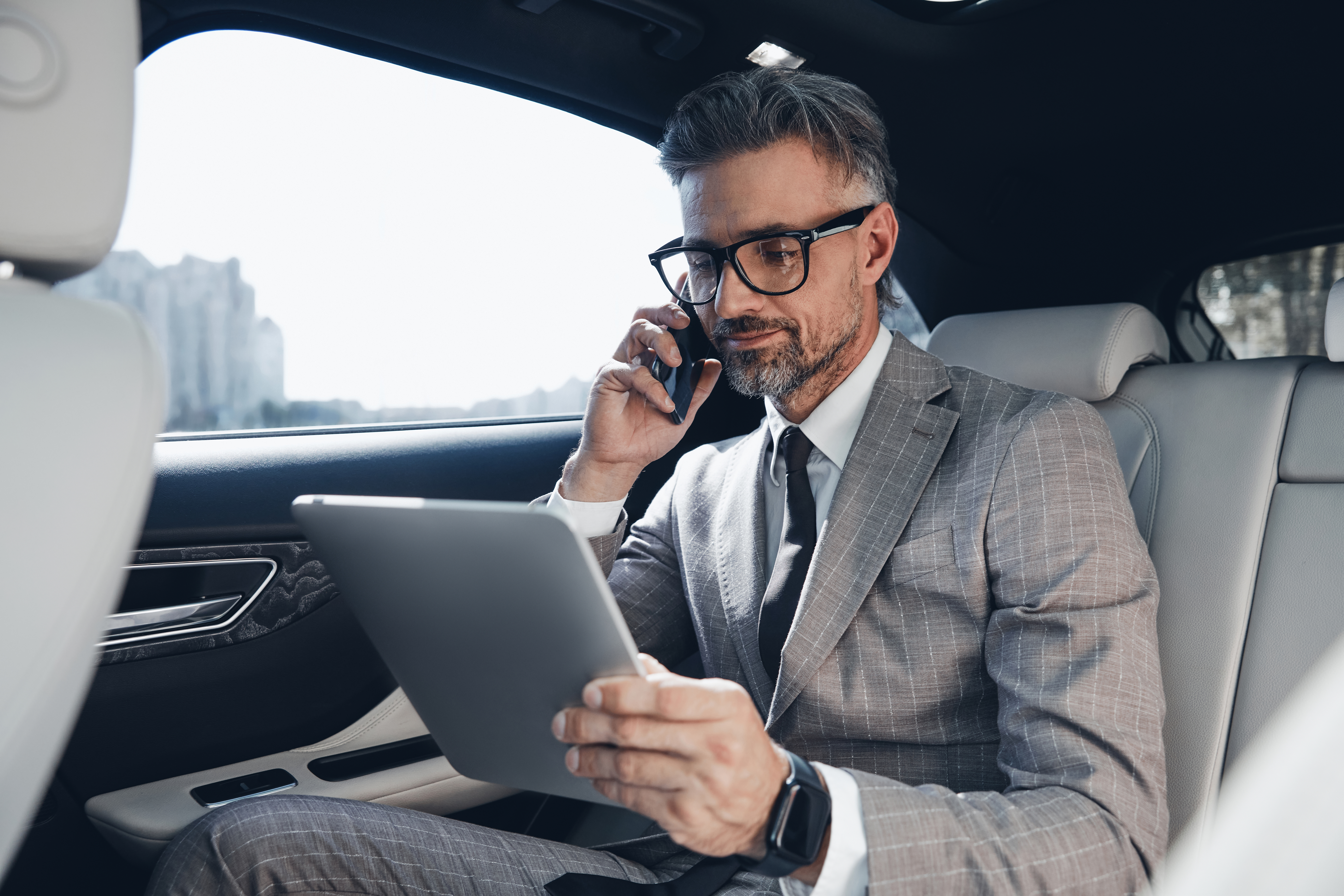 Confident businessman using digital tablet and talking on phone while sitting in the car