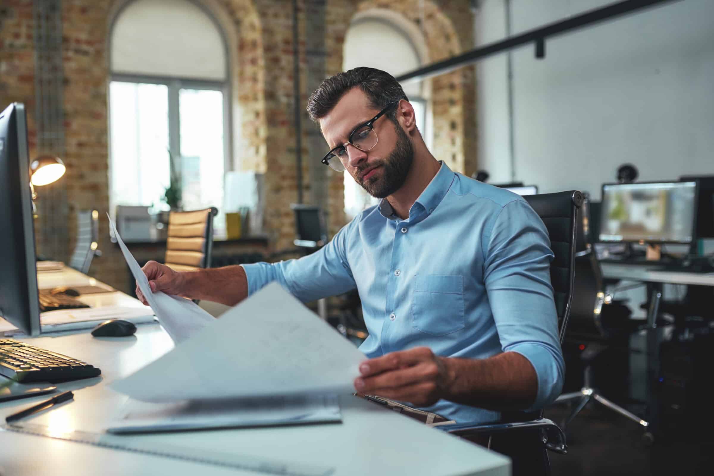 Construction concept. Concentrated bearded engineer in eyeglasses and formal wear looking at blueprint while working in the office