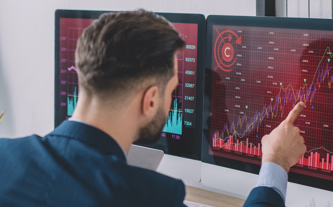 Trader pointing at market charts on two monitors