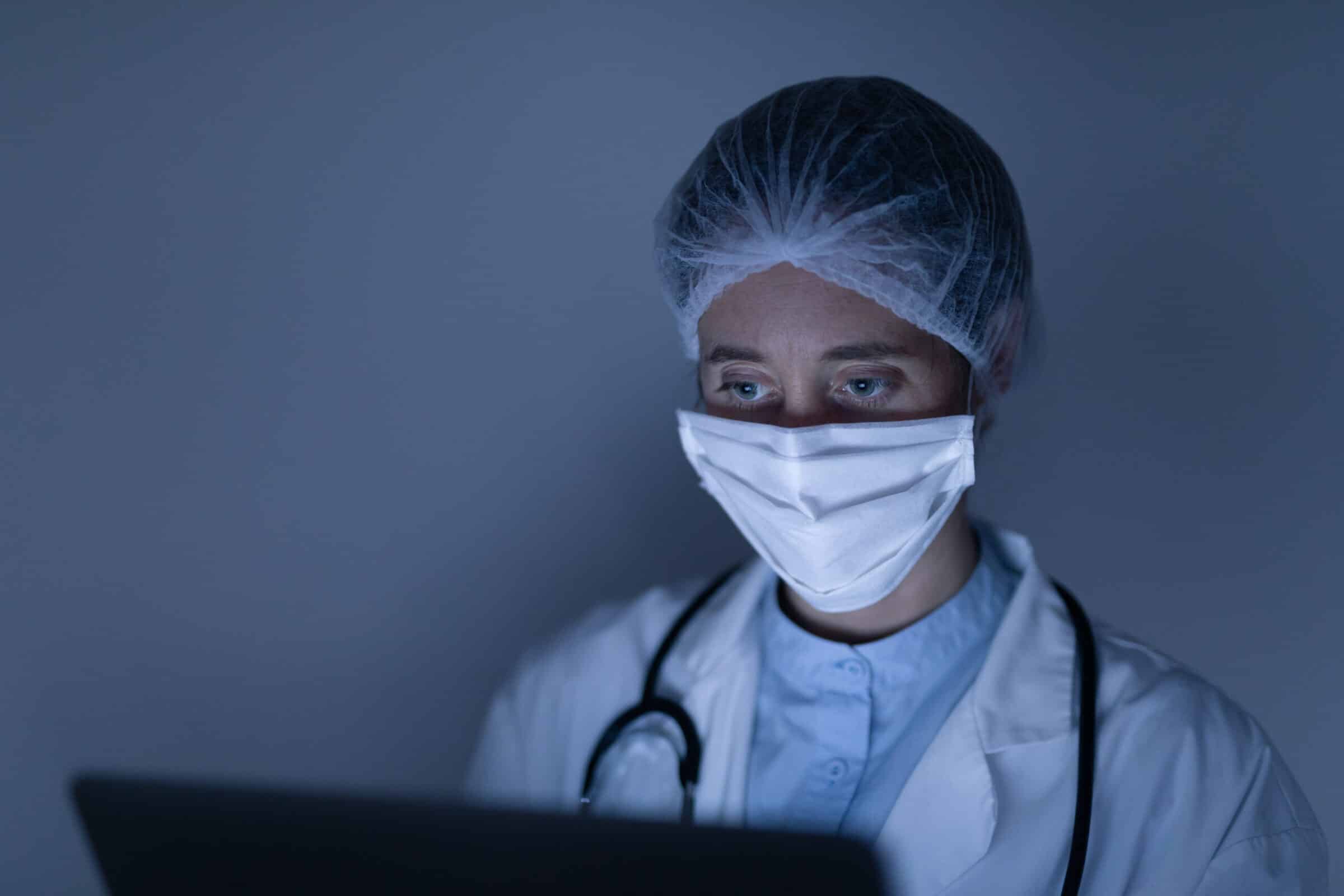 Caucasian female healthcare worker wearing a lab coat, face mask and protective cap against coronavirus, covid 19, with a stethoscope on her neck, using her laptop computer.